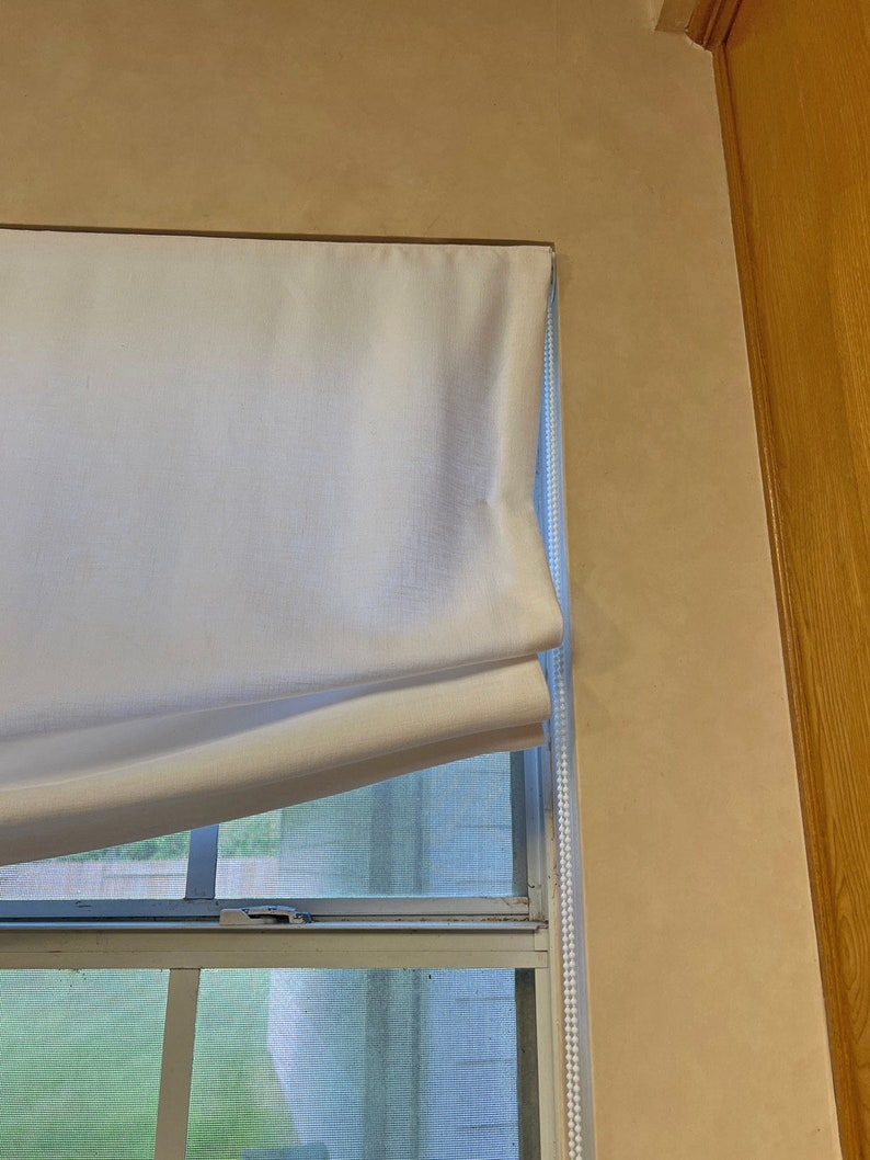 May include: A white roman shade with a white cord is pulled up to reveal a window with a white frame and a screen. The window is partially obscured by a green leafy plant outside.