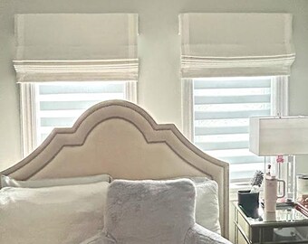 May include: Bedroom scene featuring a bed with a large, ornate headboard. A gray, plush backrest pillow sits on the bed, with white pillows behind it. Two windows with white Roman shades are visible, along with a nightstand and lamp.