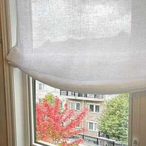 May include: White sheer linen Roman blind with a gathered valance, hanging in a window with a view of a red-leafed tree and a brick building in the background.