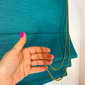 May include: Teal-colored Roman shade with a textured fabric. A gold-colored beaded chain hangs down the side. The shade is partially raised, revealing the folded design. The image shows a close-up of the shade and chain.