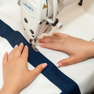May include: A person's hands are guiding a white sewing machine as it sews a blue stripe onto a white fabric.