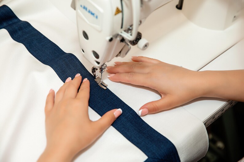May include: A person's hands are guiding a white sewing machine as it sews a blue stripe onto a white fabric.