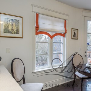 May include: White roman shade with a scalloped bottom edge and orange trim. The shade is raised, revealing a window with a view of trees and a deck. The window is framed in white trim.