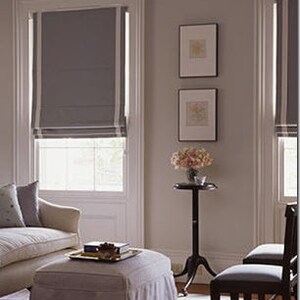 May include: A living room with a beige sofa, a white ottoman, and two chairs. The room has a window with a gray roman shade and two framed pictures on the wall. There is a small table with a vase of flowers next to the window.