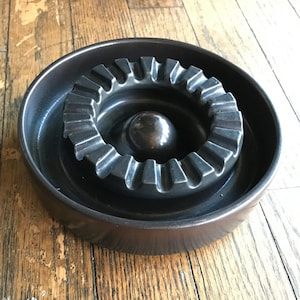 Puede incluir: Un cenicero de cerámica marrón oscuro con un anillo interior acanalado y un centro elevado. El cenicero tiene un aspecto vintage y es perfecto para contener cigarrillos.