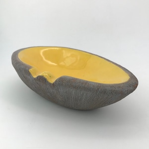 May include: Oval-shaped ashtray with a textured, dark gray exterior and a bright yellow interior. The ashtray has a small notch for resting cigarettes. The design is simple and modern, suitable for home decor.