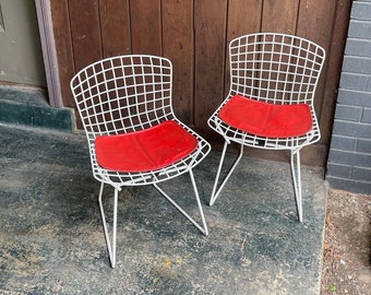 May include: Two white wire chairs with red cushions. The chairs are sitting on a concrete surface in front of a wooden wall.