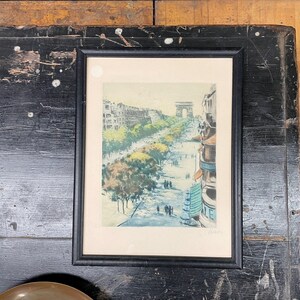 Peut inclure: Une estampe encadrée représentant une scène de rue parisienne, avec l'Arc de Triomphe en arrière-plan. L'œuvre d'art utilise une palette de couleurs sourdes avec des nuances de bleu, de vert et de brun. L'estampe est dans un cadre noir.