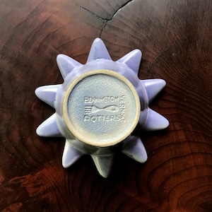 May include: A light purple ceramic dish with a starburst design. The dish has a central circular indentation and pointed edges. The bottom of the dish features the text "EDGINGTON POTTERS VERMONT" and a fish illustration.