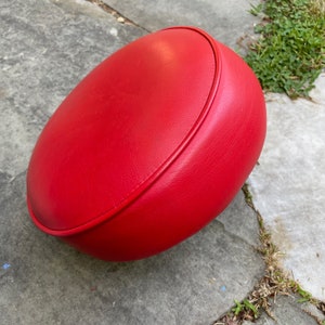 May include: A bright red round seat cushion with a smooth, slightly textured surface. The cushion is made of a soft, padded material and has a rounded edge.