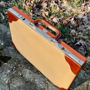 May include: A tan and brown briefcase with a brown handle and silver hardware. The corners and edges are trimmed with brown leather. The briefcase is resting on a stone surface.