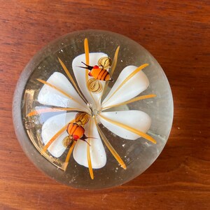 May include: A clear glass paperweight with a white flower design and orange accents. Two orange and black bee figures are inside the paperweight. The paperweight is round and sits on a wooden surface.