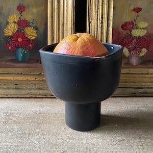 Può includere: Una ciotola in ceramica nera con un bordo largo e una base a piedistallo, che contiene un pompelmo rosa. La ciotola è appoggiata su una superficie beige testurizzata davanti a due quadri con cornici dorate con dipinti floreali.