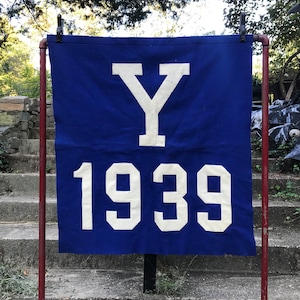 May include: A blue banner featuring a white "Y" and the year "1939". The banner is displayed on a red metal frame outdoors. The background shows a stone staircase and trees, typical of an outdoor setting.