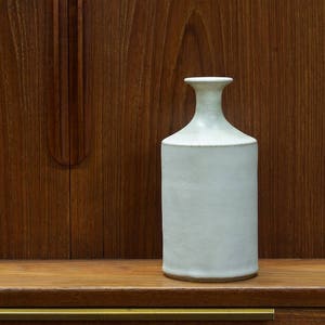 Puede incluir: Un jarrón de cerámica blanco con cuerpo redondeado y cuello estrecho. El jarrón está sobre una superficie de madera.