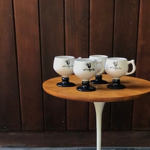 May include: Set of four white ceramic mugs with black rooster designs and black bases, displayed on a round wooden table with a white tulip-shaped base.