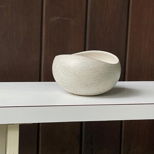 May include: A textured, off-white ceramic bowl with a unique, asymmetrical design. The bowl has a rounded shape with a curved opening, revealing a smooth, white interior. The exterior features a subtle, speckled pattern.