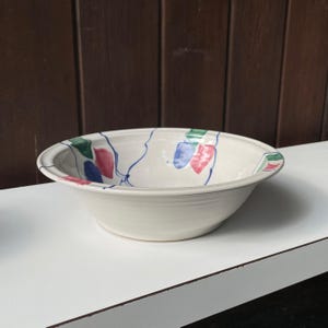 Puede incluir: Un bol de cerámica blanca con un diseño abstracto en azul, verde y rojo. El bol está sobre una superficie blanca.