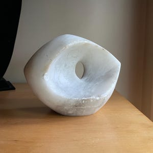 May include: A white marble sculpture with a central circular opening. The abstract form has a smooth, curved surface and a polished appearance. The sculpture is displayed on a wooden surface.