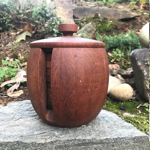 Puede incluir: Un portavasos de madera con tapa. El portavasos es de madera oscura y tiene forma redonda. La tapa también es de madera y tiene un pomo en la parte superior.