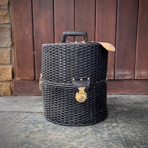 Puede incluir: Un baúl de almacenamiento de mimbre negro con una cerradura y un asa de latón. El baúl tiene un patrón tejido y está cerrado.