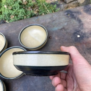 May include: A set of small ceramic bowls. Each bowl has a cream-colored interior and a dark blue band around the base. The rim is also dark blue. The bowls are displayed on a weathered surface.