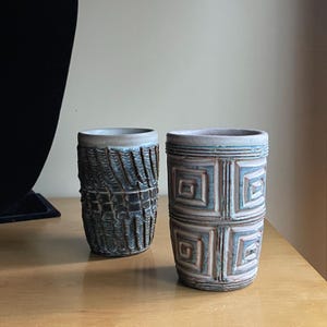 May include: Two ceramic tumblers with textured surfaces. One is dark blue with a woven pattern, the other is light blue with a geometric square design. Both have a light blue rim and sit on a wooden surface.