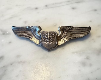 May include: A vintage silver-toned metal wing pin with a shield in the center. The wings are detailed with feather-like textures, and the shield features a detailed emblem. The pin is likely a military or aviation insignia.
