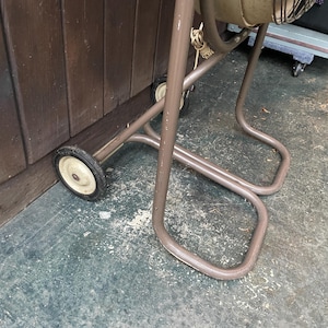 Peut inclure: Une chaise pliante en m&eacute;tal marron avec deux roues sur les pieds avant. La chaise est pli&eacute;e et appuy&eacute;e contre un mur en bois.