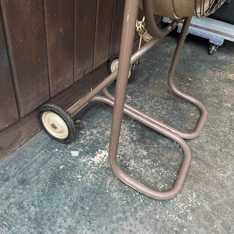 Peut inclure: Une chaise pliante en m&eacute;tal marron avec deux roues sur les pieds avant. La chaise est pli&eacute;e et appuy&eacute;e contre un mur en bois.