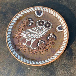 May include: A decorative ceramic plate featuring a detailed owl design. The plate has a brown and white color scheme with a textured surface. The owl is depicted in white with brown accents, surrounded by floral patterns.