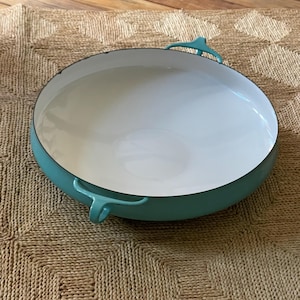 Puede incluir: Una sartén de hierro fundido esmaltado turquesa con interior blanco. La sartén tiene dos asas turquesas y un borde negro. La sartén está sobre una superficie tejida de color beige.