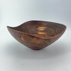 May include: A dark brown wooden bowl with a unique, abstract design carved into the surface. The design is in a lighter brown color, creating a contrast against the dark wood.