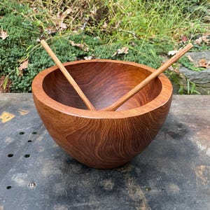 May include: A large, round wooden bowl with a rich reddish-brown color. The bowl has a smooth, polished finish and is made of a single piece of wood. Two wooden spoons are resting inside the bowl.