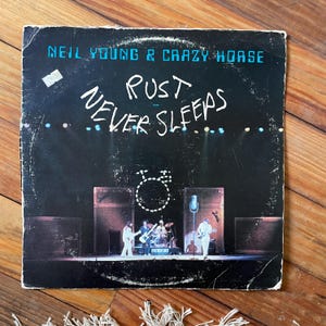 May include: Vintage vinyl record album cover for "Rust Never Sleeps" by Neil Young & Crazy Horse. The album cover features the band performing on stage, with the album title in white script and the band's name in blue.
