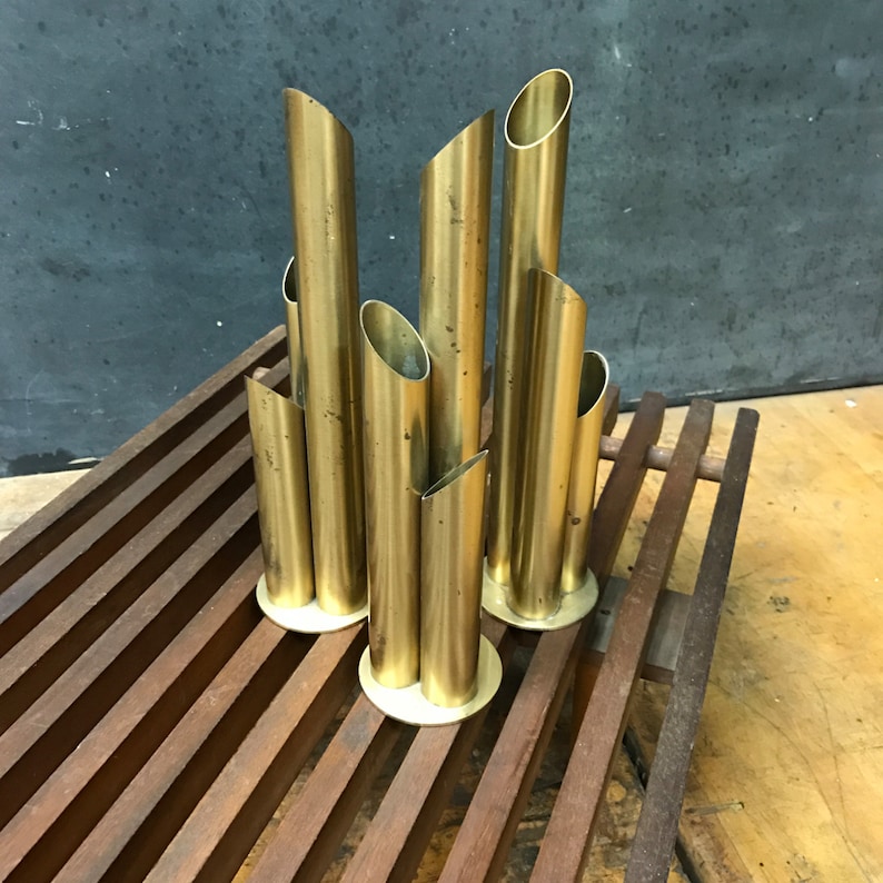 May include: A set of gold metal bud vases arranged in a staggered formation. The vases are made of polished brass and have a cylindrical shape. The vases are arranged on a wooden surface.