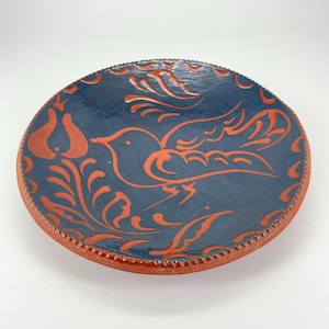 May include: A round, handmade ceramic plate with a dark blue interior and a reddish-orange rim. The plate features a bird and floral design in reddish-orange. The rim has a decorative, scalloped edge. The plate is likely used for serving food or as a decorative piece.
