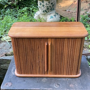 May include: A rectangular wooden storage box featuring a smooth top surface and two vertical tambour doors that slide open. The unit is constructed from medium-toned wood with a visible grain pattern and includes two integrated vertical wooden handles.