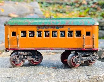 May include: A vintage toy train car painted in orange and green with the word "PULLMAN" printed on the side. The car has windows with painted figures inside and four wheels.
