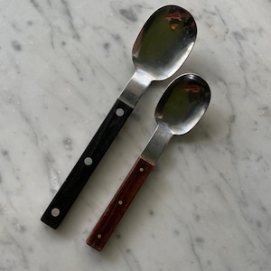 May include: Two metal spoons with wooden handles. The larger spoon has a black handle and the smaller spoon has a brown handle.