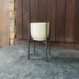 May include: White ceramic planter on a black metal stand. The stand has four legs and is designed to elevate the planter.