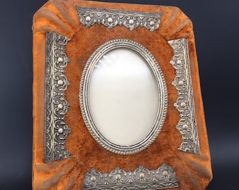 May include: An antique photo frame with an oval opening. The frame is covered in orange velvet and has ornate silver-colored metal trim. The frame is rectangular with rounded corners, and the velvet is slightly gathered around the edges.