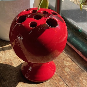 May include: A red ceramic flower vase with a round, spherical body and a short, round base. The vase has multiple holes around the top for holding flower stems.