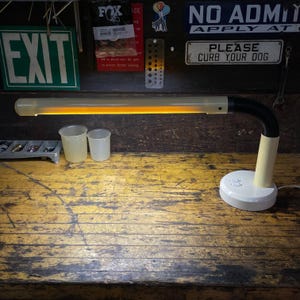May include: A white desk lamp with a long, thin, orange fluorescent light tube. The lamp is on a wooden table with two clear plastic cups.
