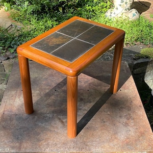 May include: A rectangular side table with a light brown wooden frame and four cylindrical legs. The tabletop features four inset square slate tiles in a dark gray and charcoal color palette with natural variations in texture and tone.