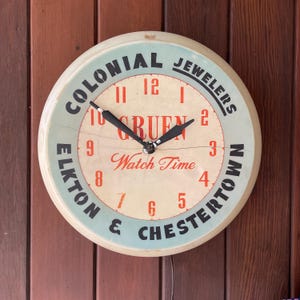 May include: A white circular clock with black hands and black lettering. The clock face reads "Colonial Jewelers Elkton & Chestertown" and "Gruen Watch Time".