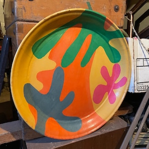 May include: A round, colorful tray with a bright orange, yellow, green, and blue abstract design. The tray is made of metal and has a slightly raised rim.