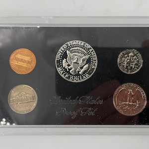 May include: A United States Proof Set in a clear plastic case. The set includes a penny, nickel, dime, quarter, and half dollar. The coins are displayed on a black background, with the words "United States Proof Set" printed below.