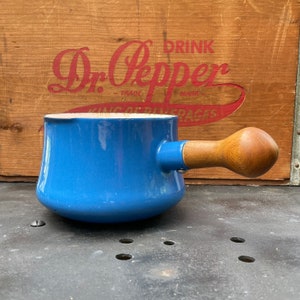 Puede incluir: Una cacerola de esmalte azul con un mango de madera. La cacerola está sobre una superficie negra.