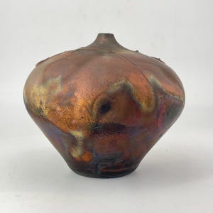 May include: A rounded ceramic vase with a narrow neck and a wide, bulbous body. The surface features a metallic, iridescent glaze with blended tones of copper, bronze, dark brown, and hints of gold, creating a swirling, multi-colored finish.